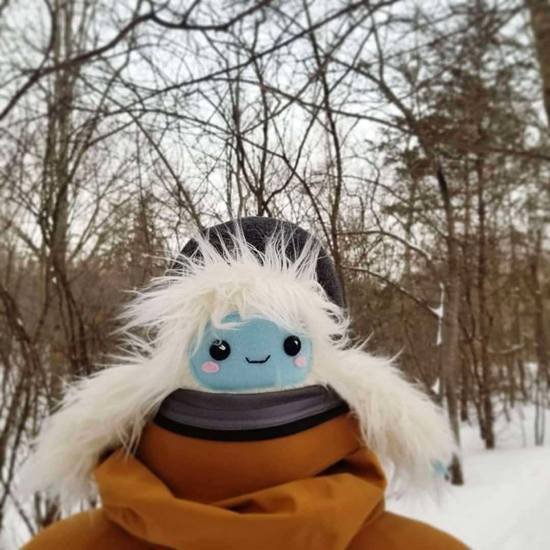 Fuzzy Yeti plush toy sitting in person's coat hood outside in the snow